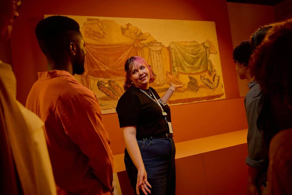 A museum guide explains a wall painting to visitors in a gallery with warm lighting and orange walls.