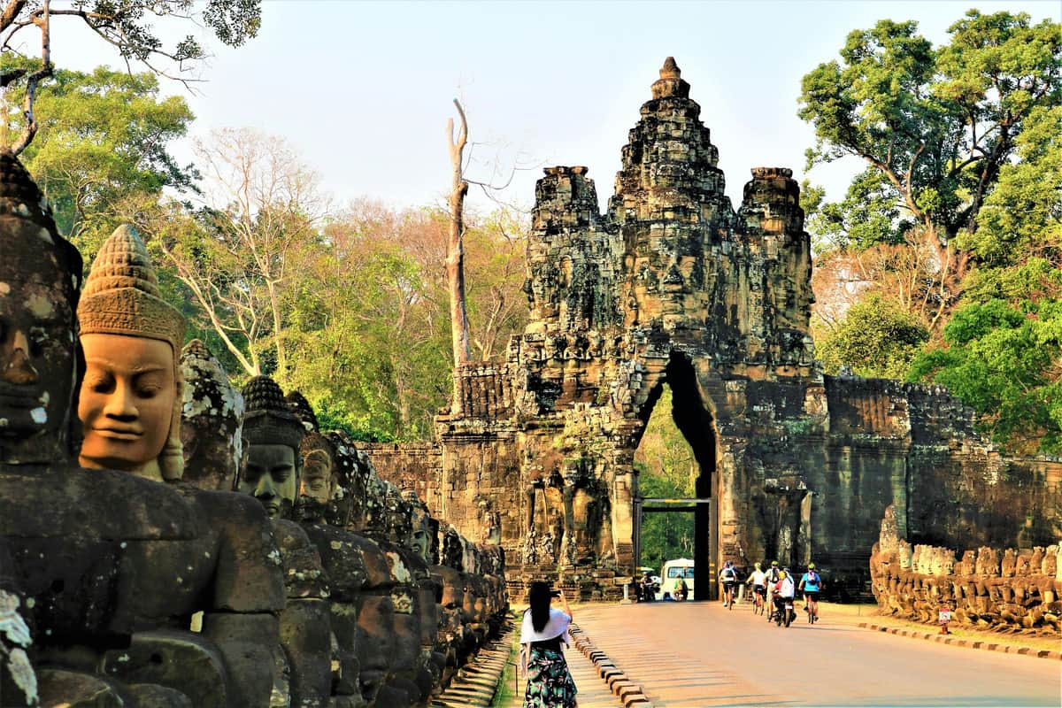 A line of stone Buddha statues leads to an ancient stone gateway; a woman in white and black stands near the entrance.