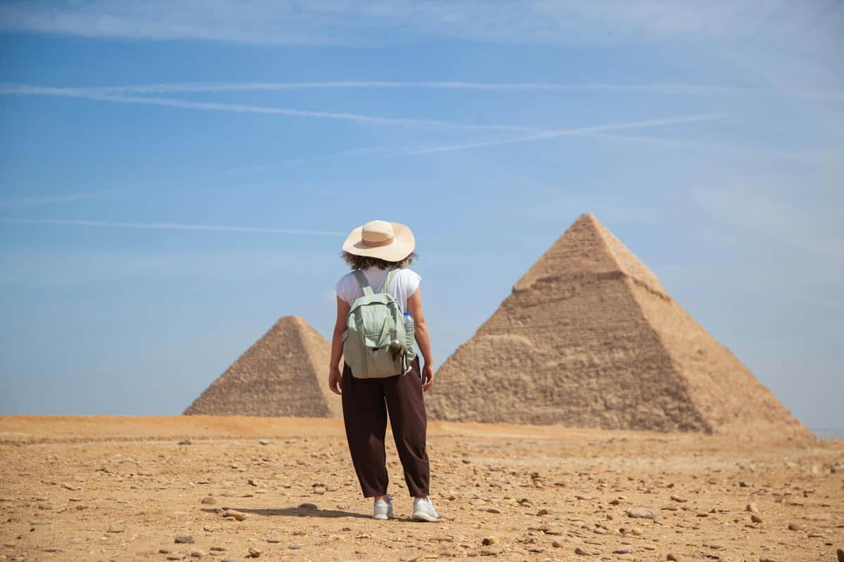 Giza Pyramids, Sphinx & Egyptian Museum: Guided Tour from Hurghada A person wearing a hat and backpack stands in front of two pyramids under a blue sky.