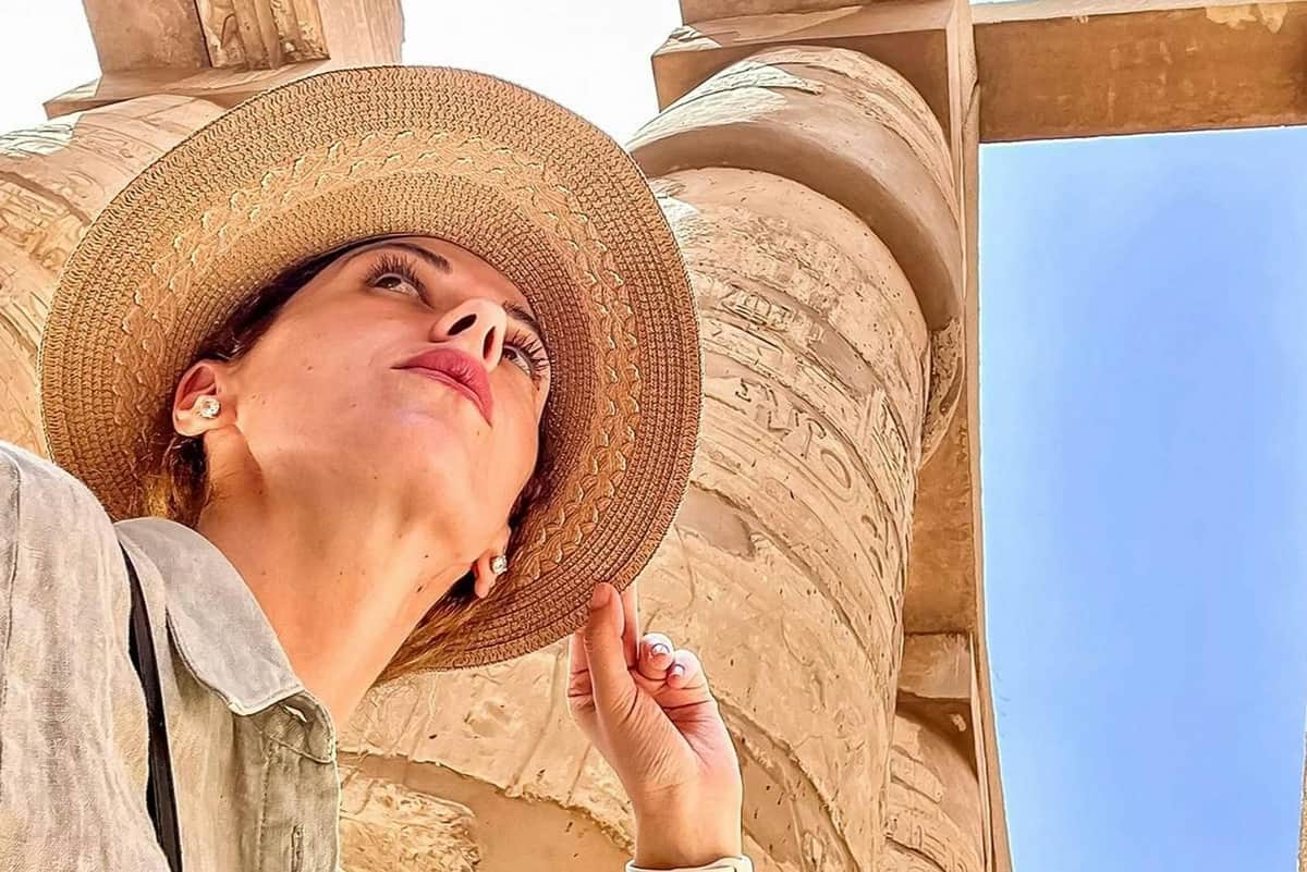 Person wearing a wide-brimmed hat, looking up at ancient stone pillars with hieroglyphics, under a bright sky.