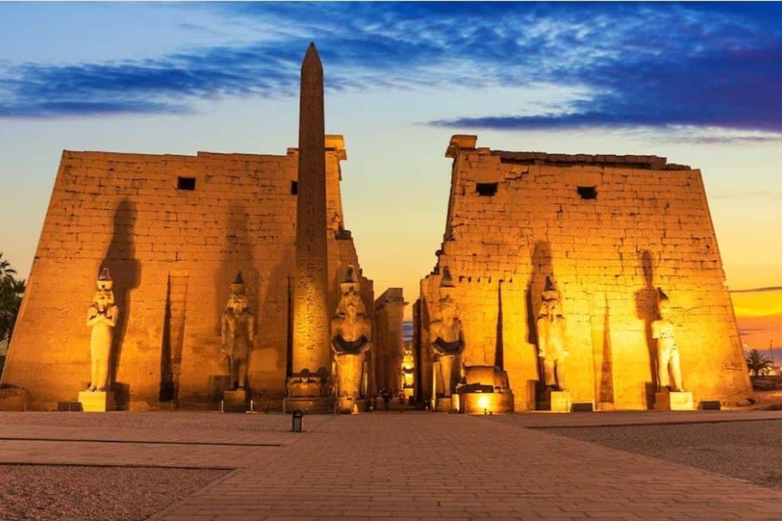 An illuminated ancient Egyptian temple facade at dusk, with statues, an obelisk, and a dramatic sky in the background.