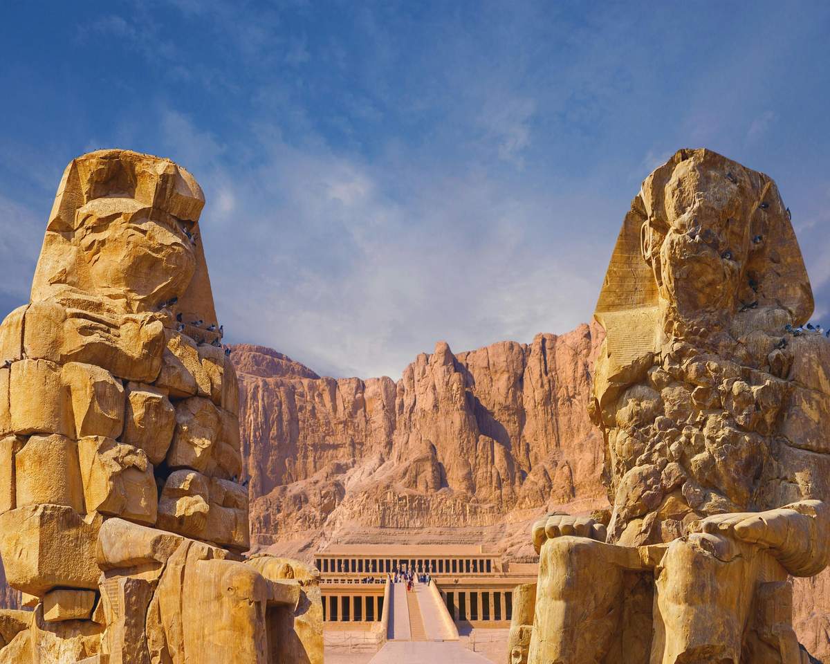 Ancient Egyptian temple entrance flanked by two large stone statues with mountainous desert landscape in the background.