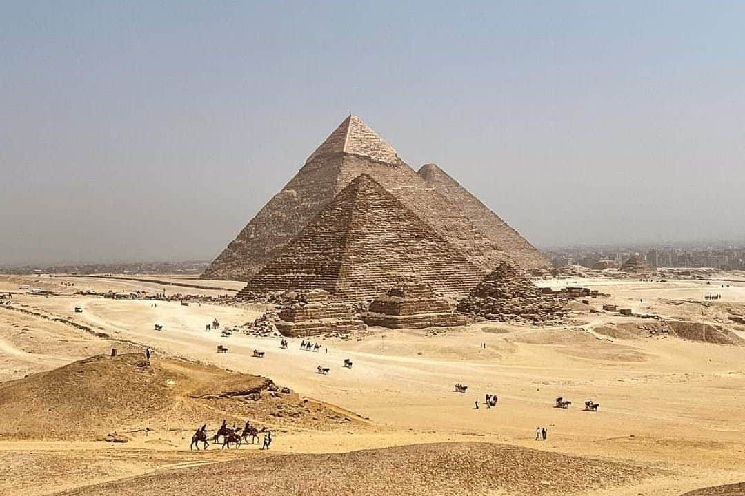 Three pyramids in a desert with people riding camels and others walking nearby under a clear sky.