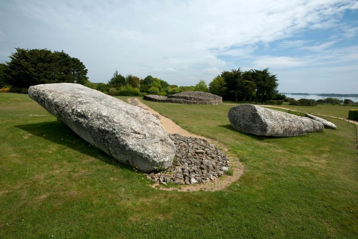Large ancient stones surrounded by grass and a gravel path, with trees and a cloudy sky in the background.