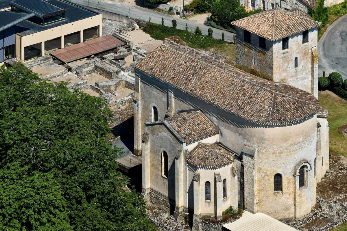 Aerial view of an old stone church with a tiled roof, adjacent ruins, and nearby buildings and greenery.