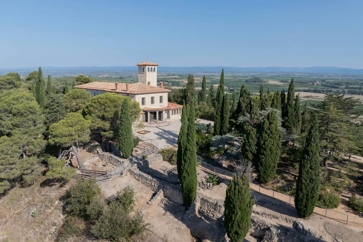 A villa with a tower surrounded by cypress trees and ruins, set in a landscape with distant fields and hills.