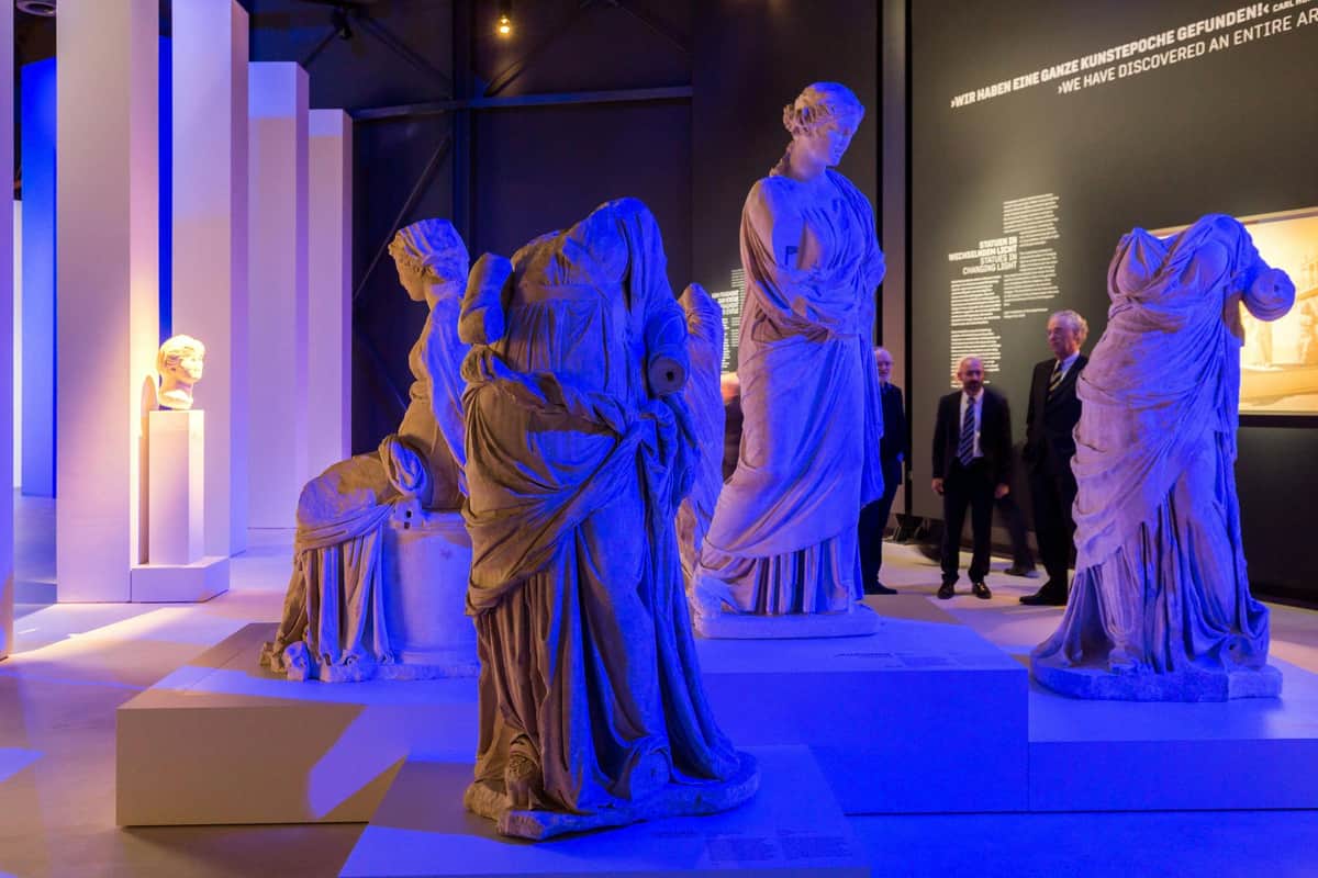 Pergamon Museum – The Panorama: Exhibition by Yadegar Asisi Exhibition of ancient statues with blue lighting, several people in suits observing, and a dark wall with text in the background.