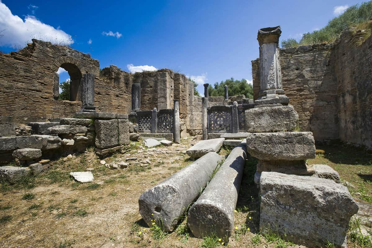 Ancient Olympia Archaeological Site & Museums: Entry Ticket Ancient stone ruins with broken columns under a clear blue sky. Surrounding arches and walls show weathered stonework.