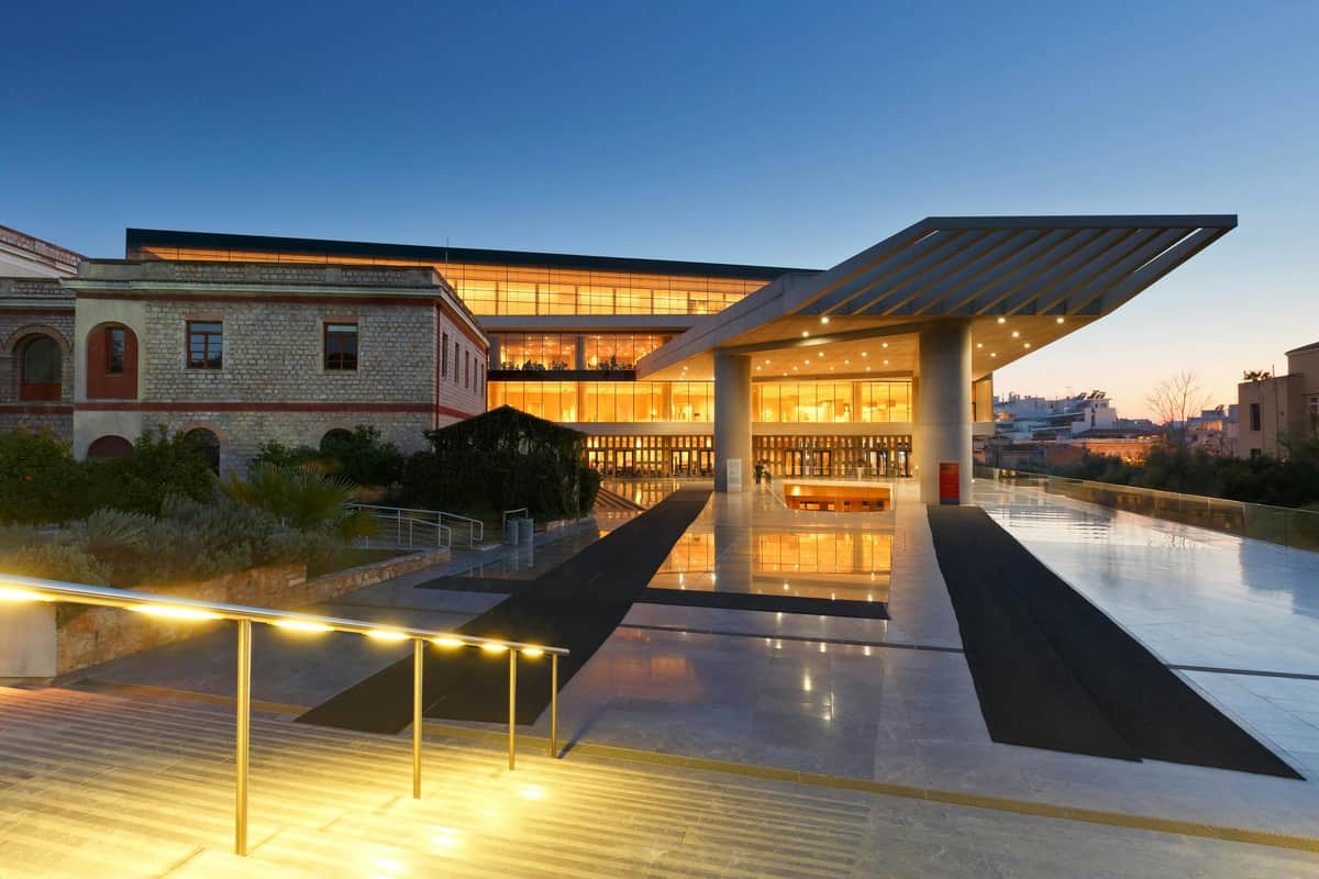 Acropolis Museum: Skip The Line Ticket + Audio Guide Modern building entrance illuminated at dusk, featuring large glass windows and reflecting surfaces, with steps and handrails in the foreground.