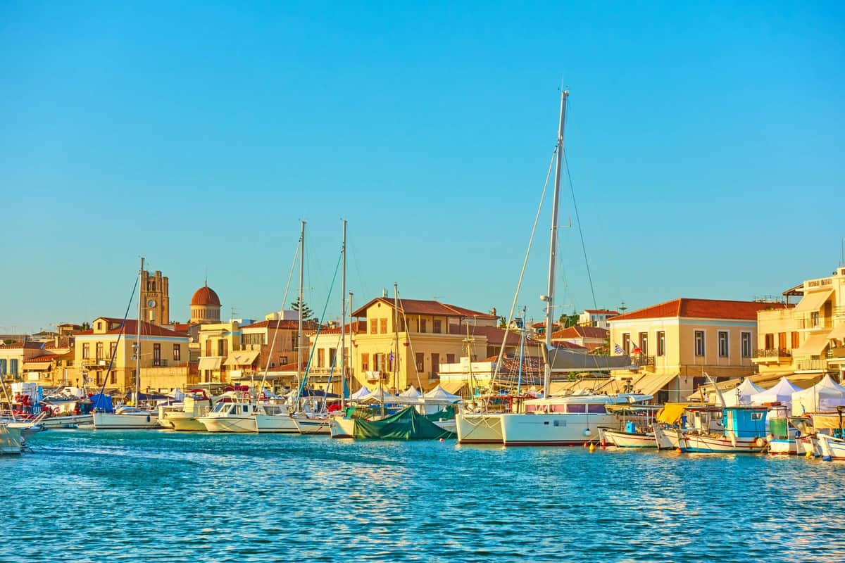 Aegina: Guided Tour from Athens + Optional Ferry Ticket Boats anchored at a harbor with a coastal town and buildings in the background under a clear blue sky.