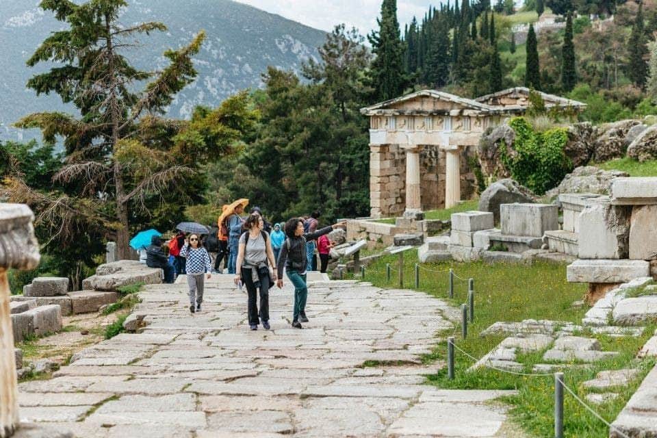 Delphi & Arachova: Day Trip from Athens People walking on a stone path with ancient ruins, trees, and mountains in the background.