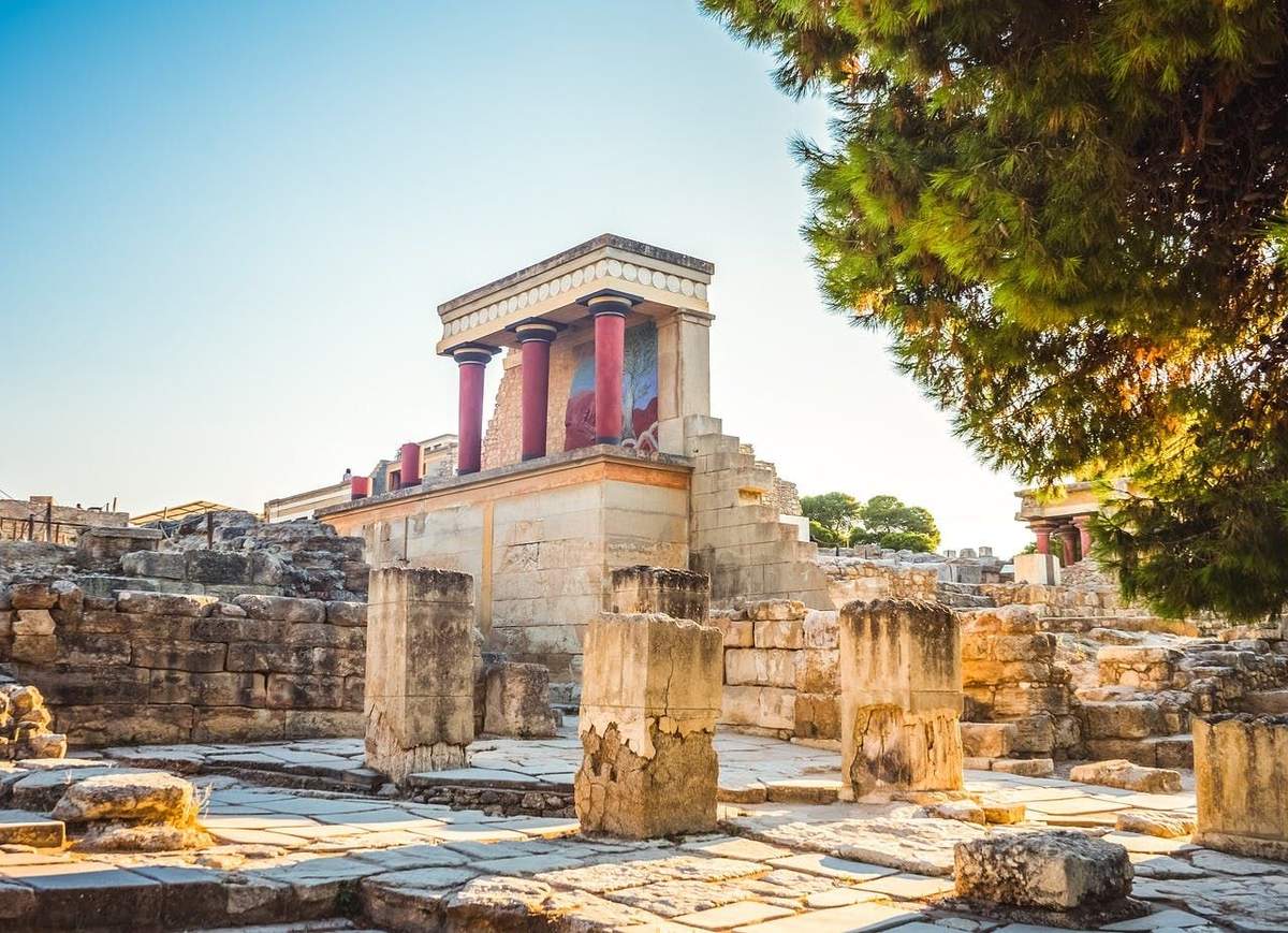 Palace of Knossos: Entry Ticket + Digital Hellenic Heritage Guide Ancient ruins with columns and a partially restored building featuring red pillars and fresco remnants under a clear blue sky.