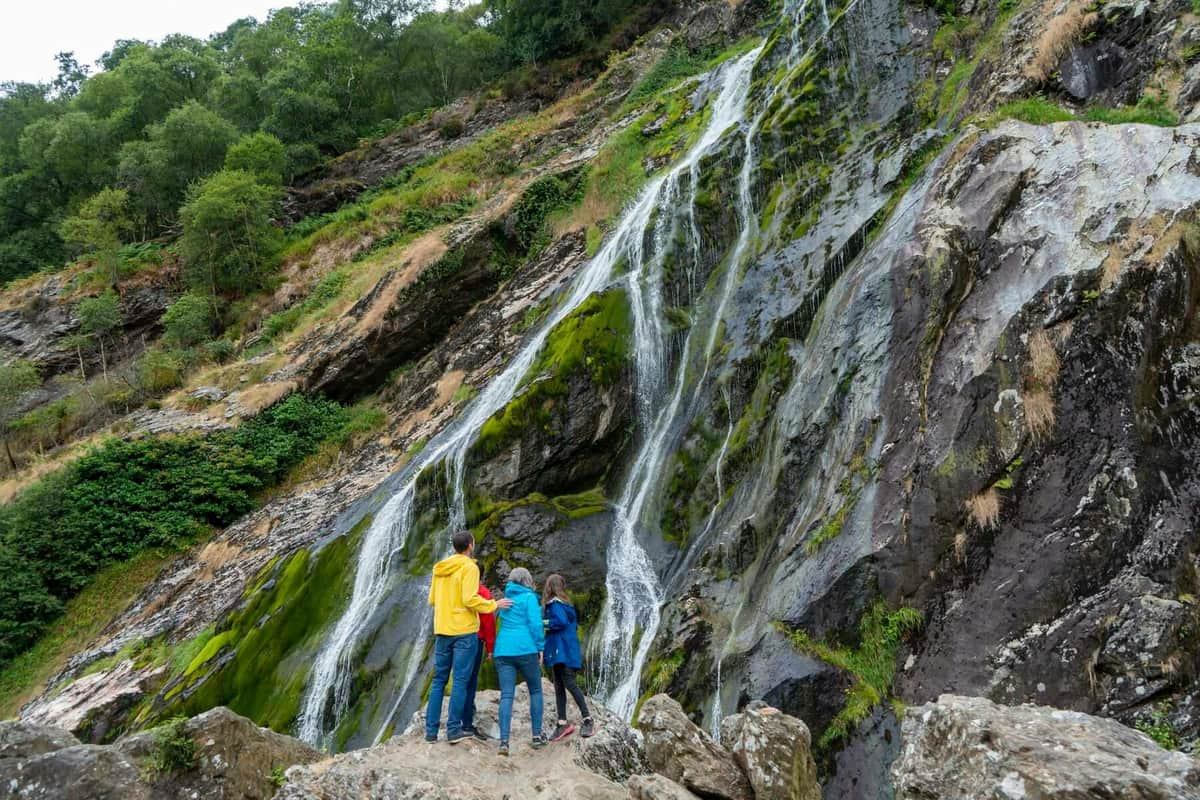 Three people in colorful jackets stand on rocks, admiring a tall, moss-covered waterfall surrounded by lush greenery.