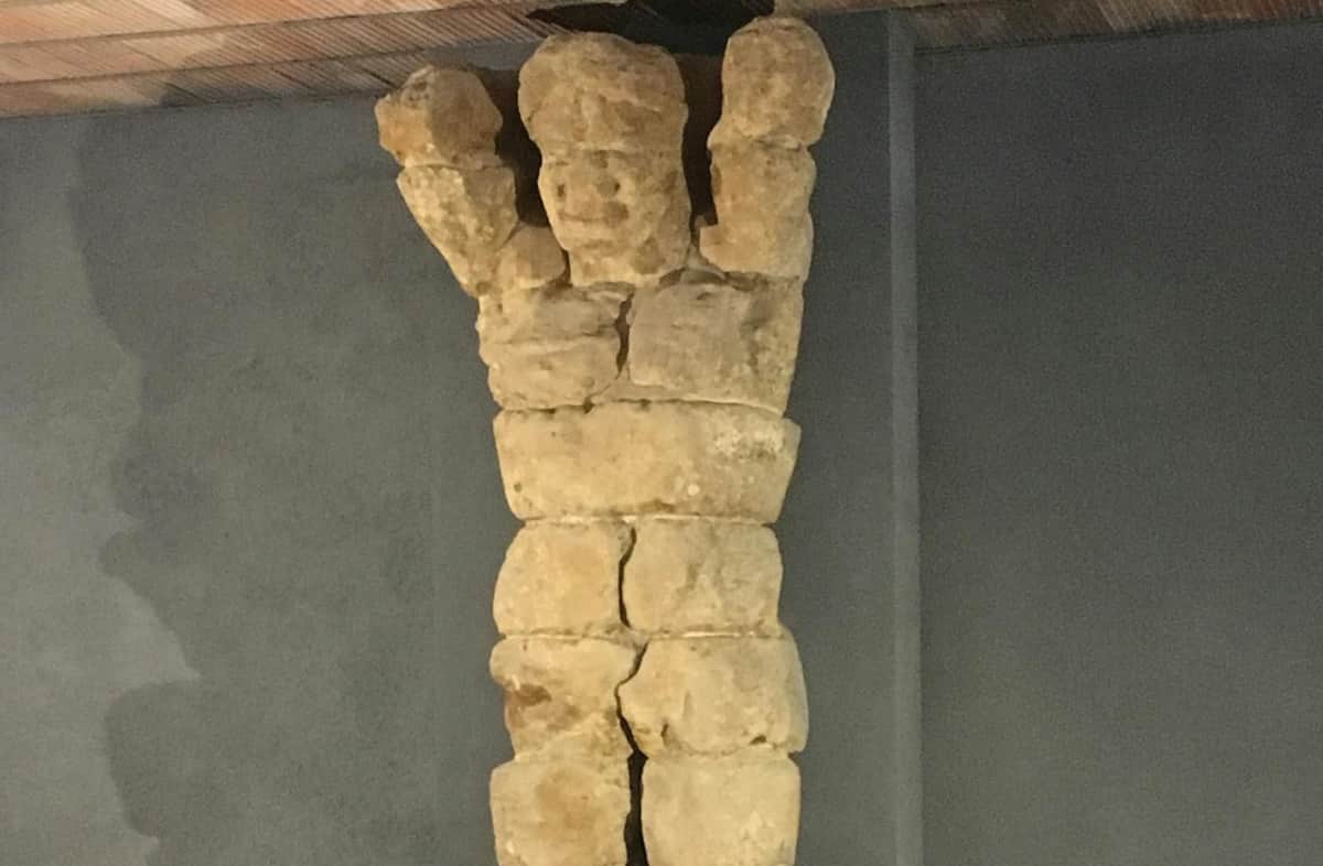 A sculpture made of stacked, irregular stone blocks resembling a human figure with raised arms against a plain wall.