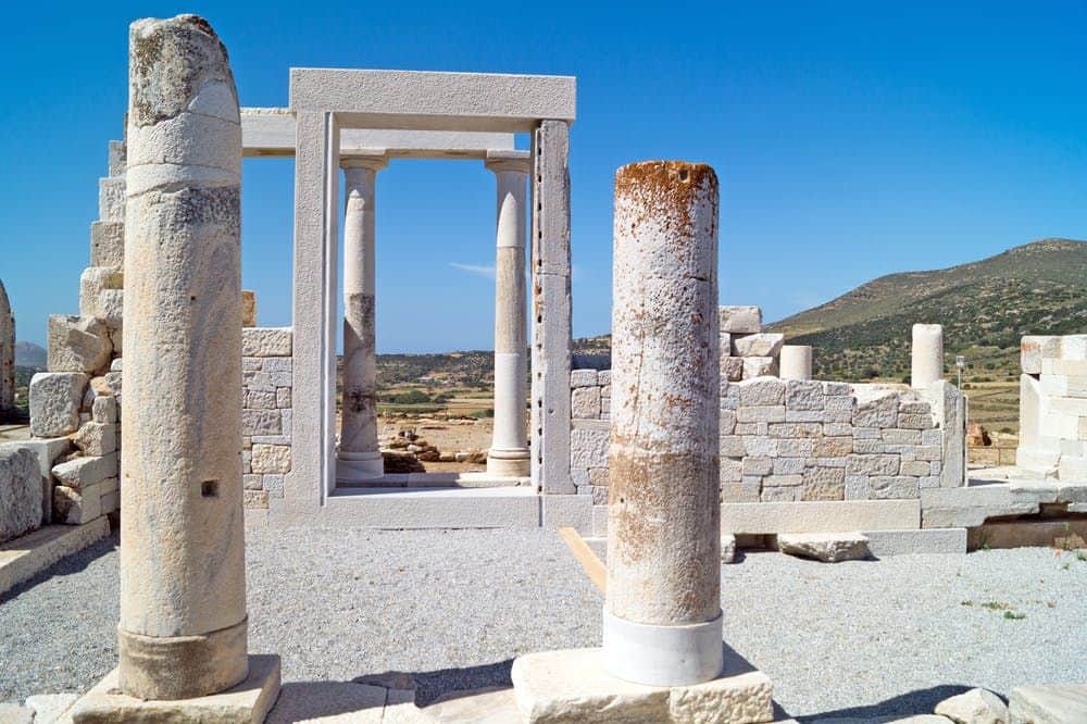 Ancient stone ruins with columns and an archway under a clear blue sky, surrounded by a rural landscape and distant hills.