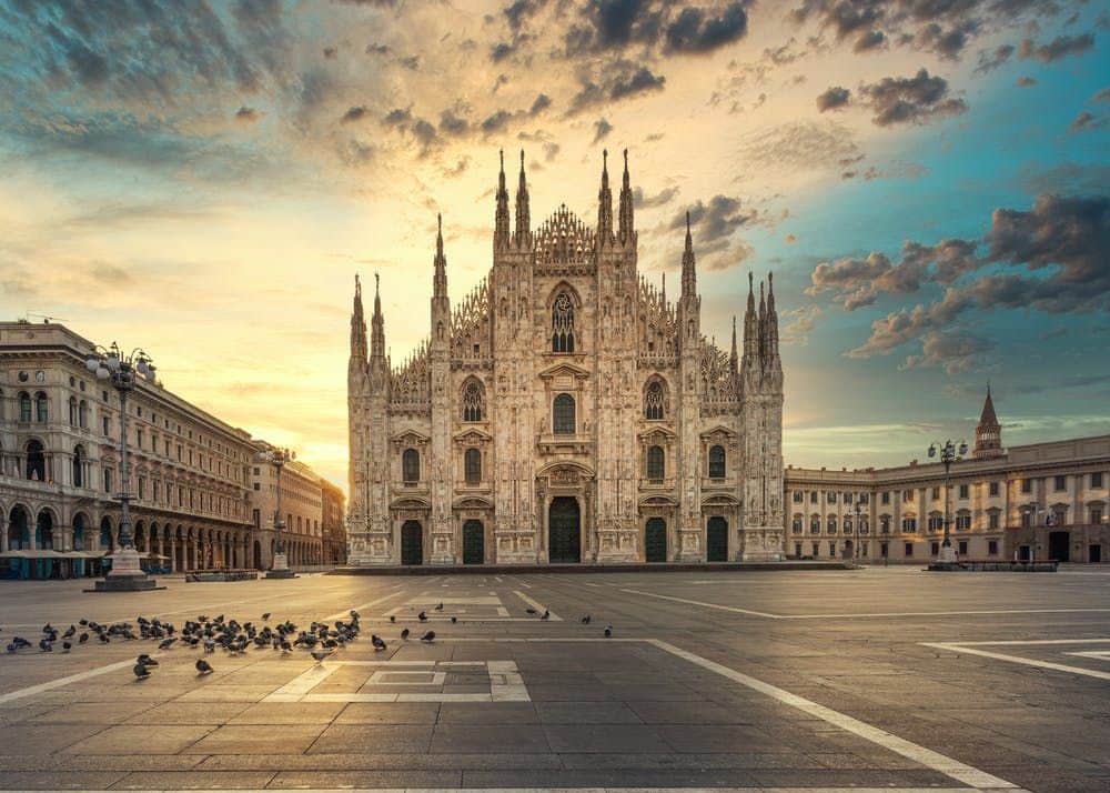Duomo di Milano: Small Group Guided Tour + Rooftop Access Piazza with scattered pigeons, large ornate cathedral with spires in background, under a dramatic sunset sky with clouds.