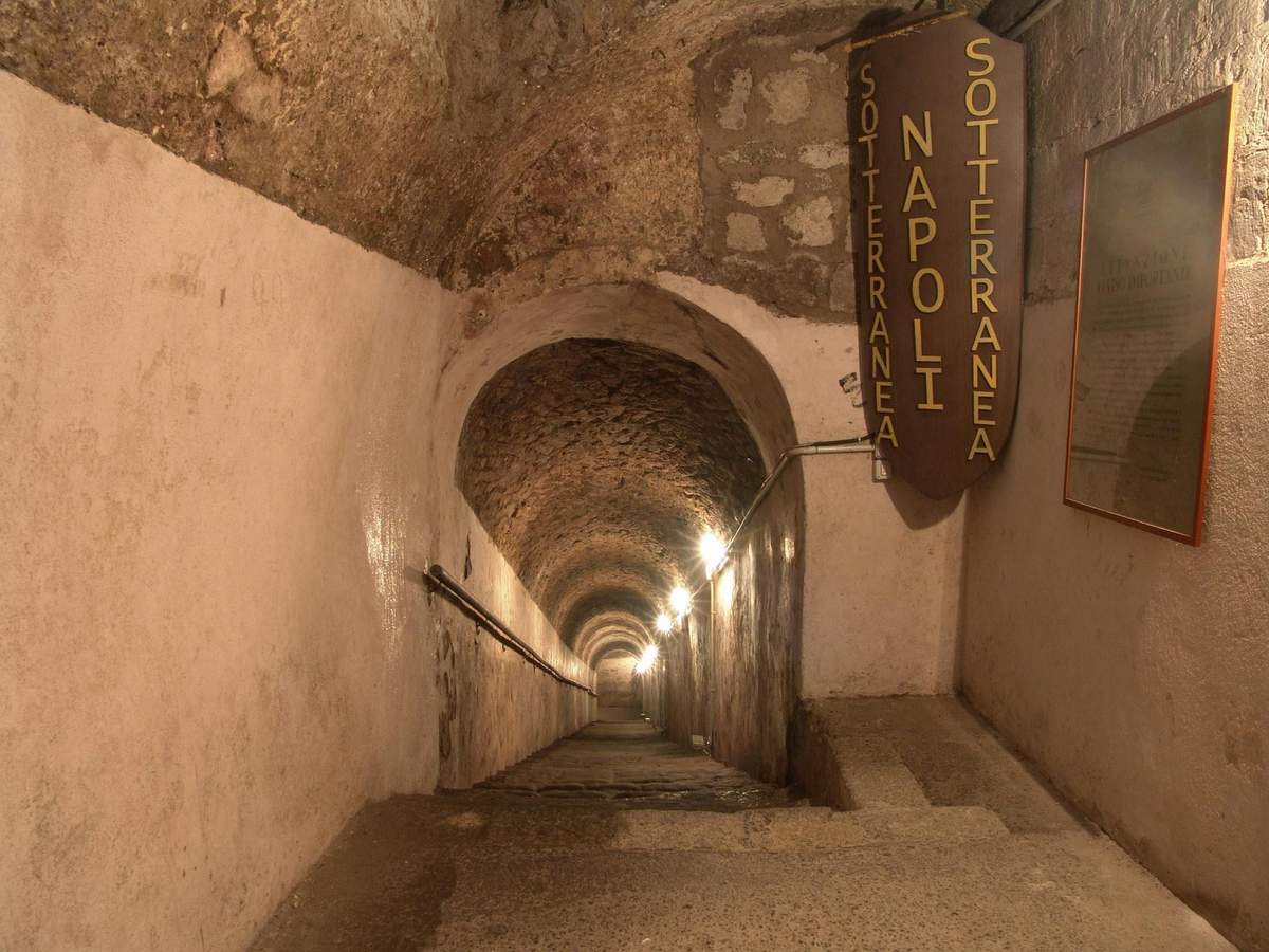 Naples Underground: Skip The Line Ticket + Guided Tour A long, narrow stone corridor with arched ceilings and wall lamps leads downwards; signs read "Napoli Sotterranea."