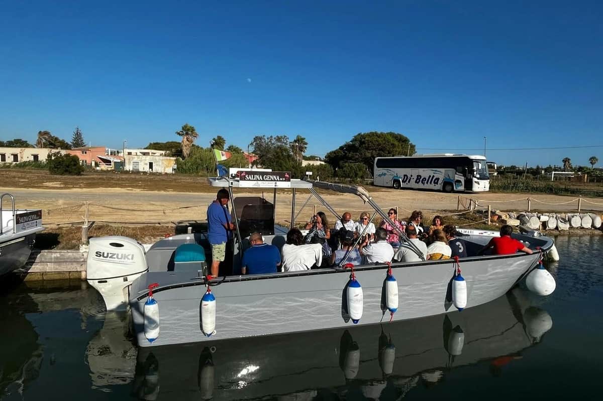 A group of people are seated on a small boat docked by a canal, with a bus and trees visible in the background under a clear blue sky.