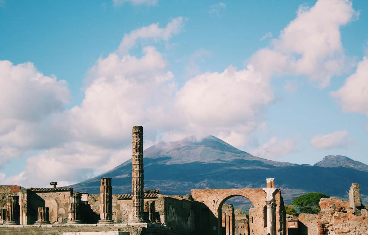 Ancient ruins with columns and arches under a blue sky, with a mountain in the background partially covered by clouds.