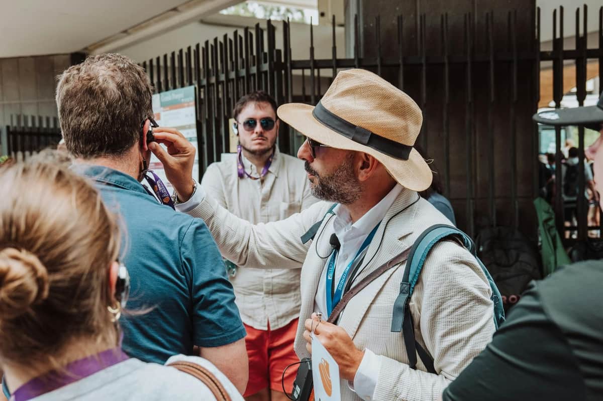 A guide in a light suit and hat adjusts a headset for a tourist in a group, with other people and a fence in the background.