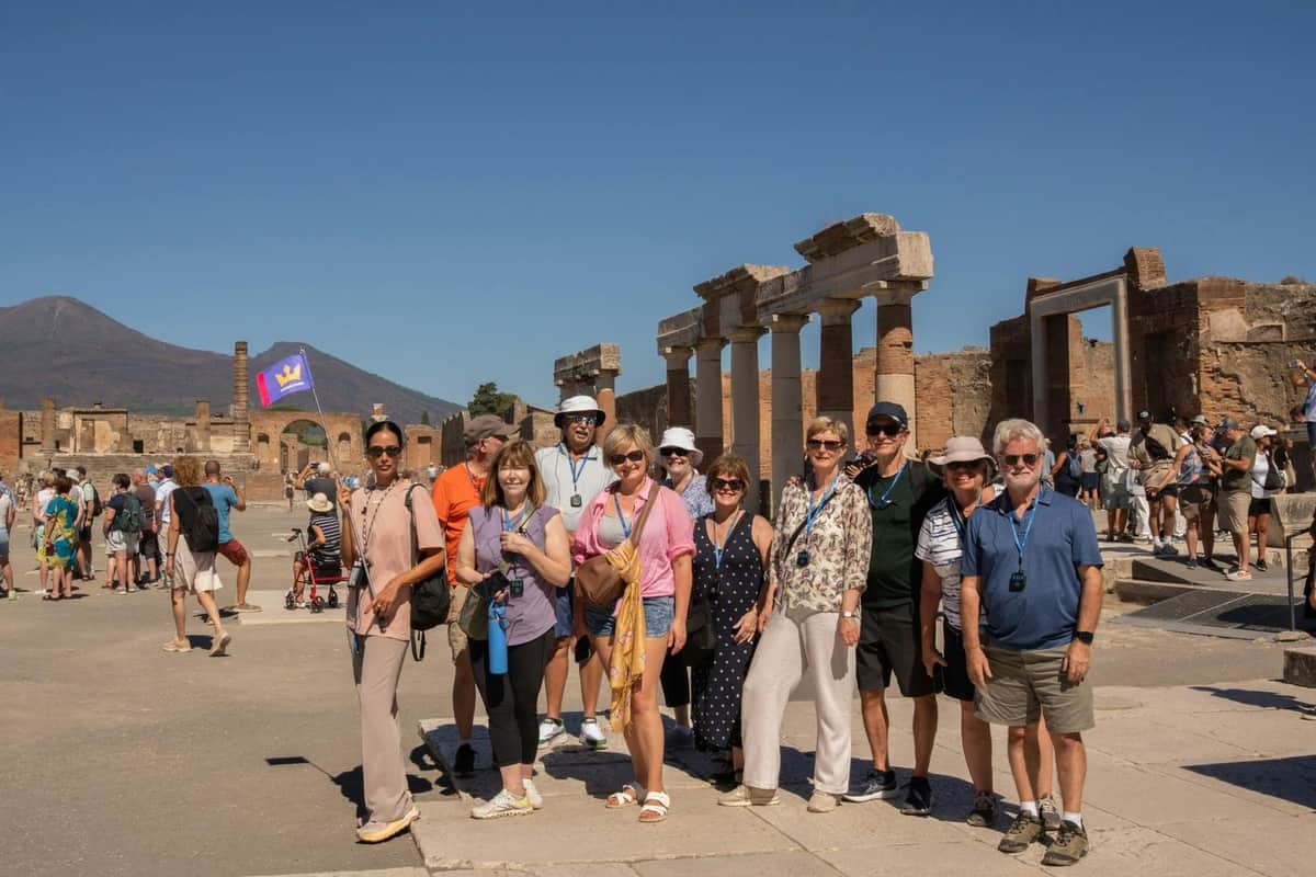 A group of tourists in front of ancient ruins with columns, under a clear blue sky. More tourists are seen in the background.