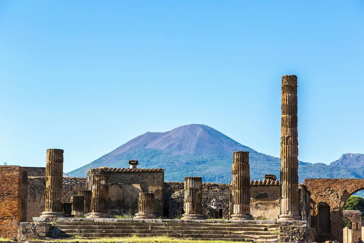 Ruins of ancient structures with tall, weathered columns in the foreground and a large mountain in the background under a clear blue sky.