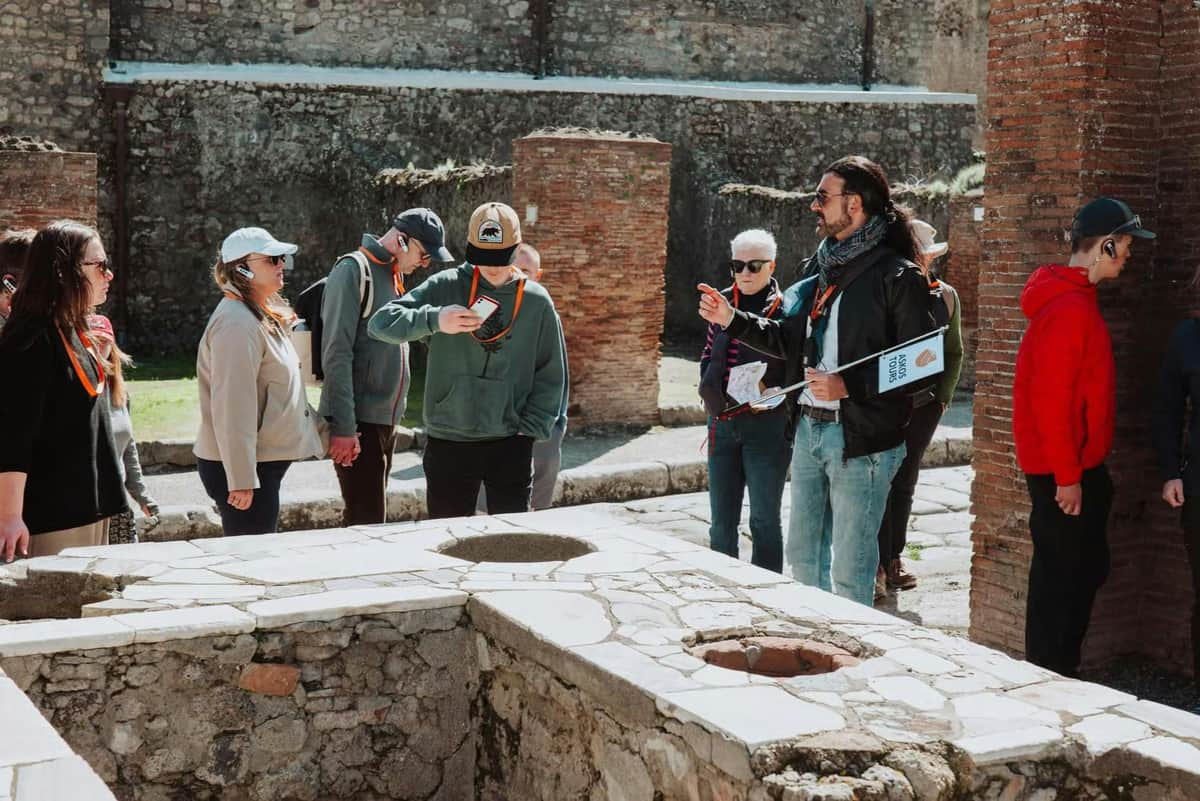 A group of people with orange lanyards stand near ancient ruins, with stone walls and grassy areas in the background.