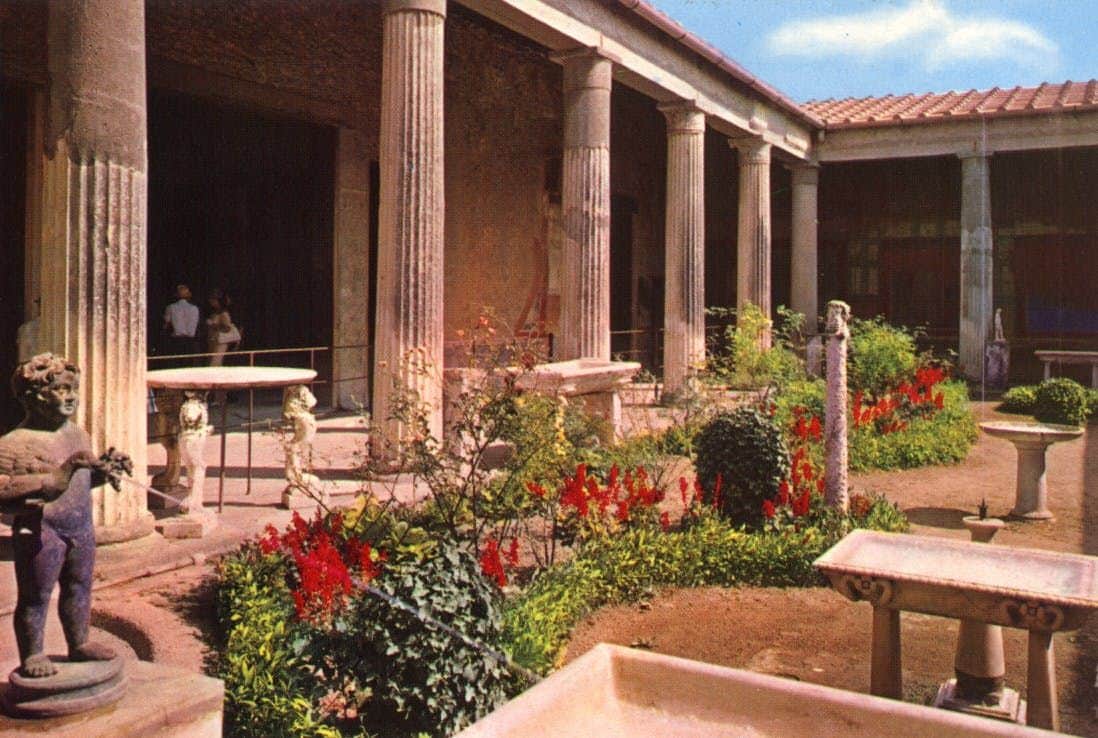 A courtyard with tall, stone columns, vibrant red flowers, and green shrubs. A few people stand in the shaded area.