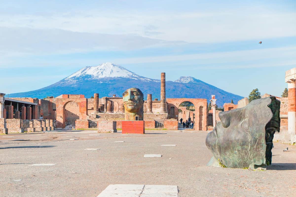 Pompeii: Skip The Line Ticket + Guided Tour Ancient ruins with large sculpted heads and Mount Vesuvius in the background under a clear sky.