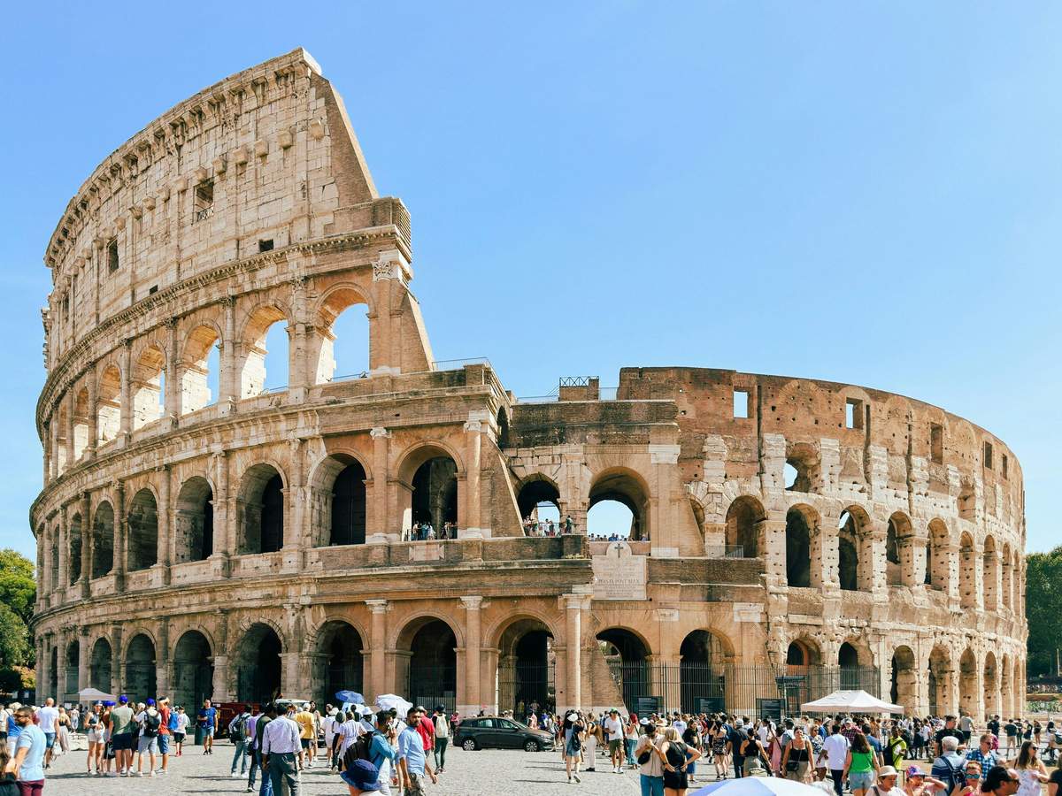 A group of tourists standing in the center of the ancient Colosseum, with its tiered stone structure and arches visible under a blue sky.