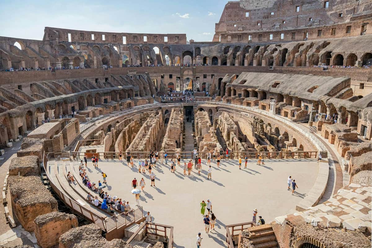 Tourists explore the interior of the Roman Colosseum with its ancient stone structure, arched openings, and exposed underground passages.