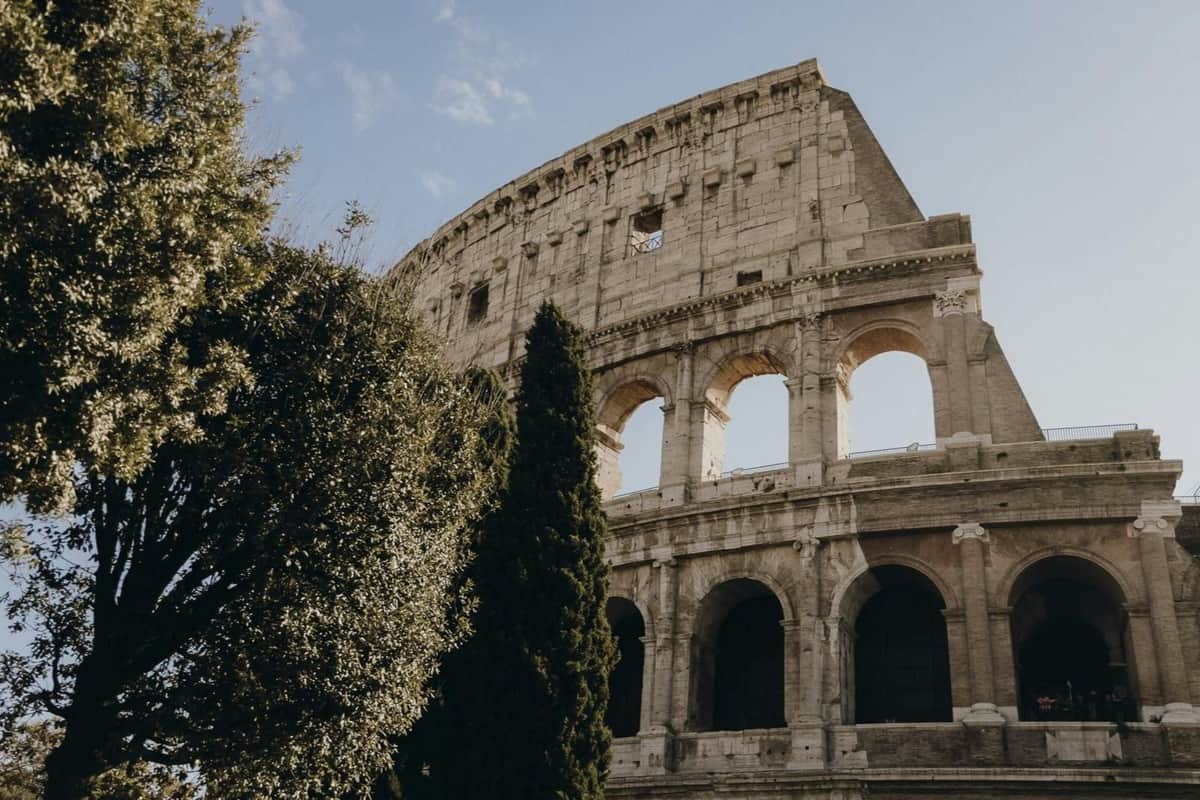 The Colosseum in Rome, partially obscured by trees, under a clear blue sky.