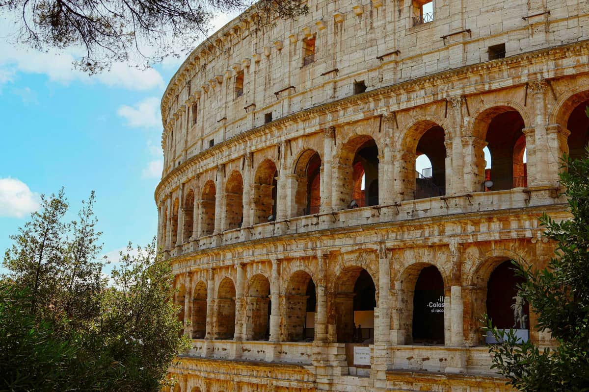 Colosseum, Roman Forum & Palatine Hill + Hop-on Hop-off Bus Tour A partial exterior view of the Roman Colosseum, showing its arched structure with trees in the foreground on a sunny day.