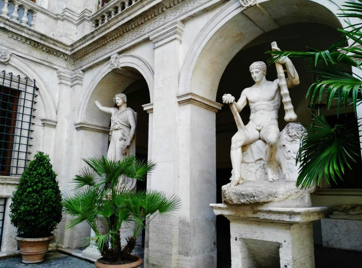 Two marble statues stand in an arched courtyard; one seated with a club, the other standing with an arm raised. Palm plants nearby.