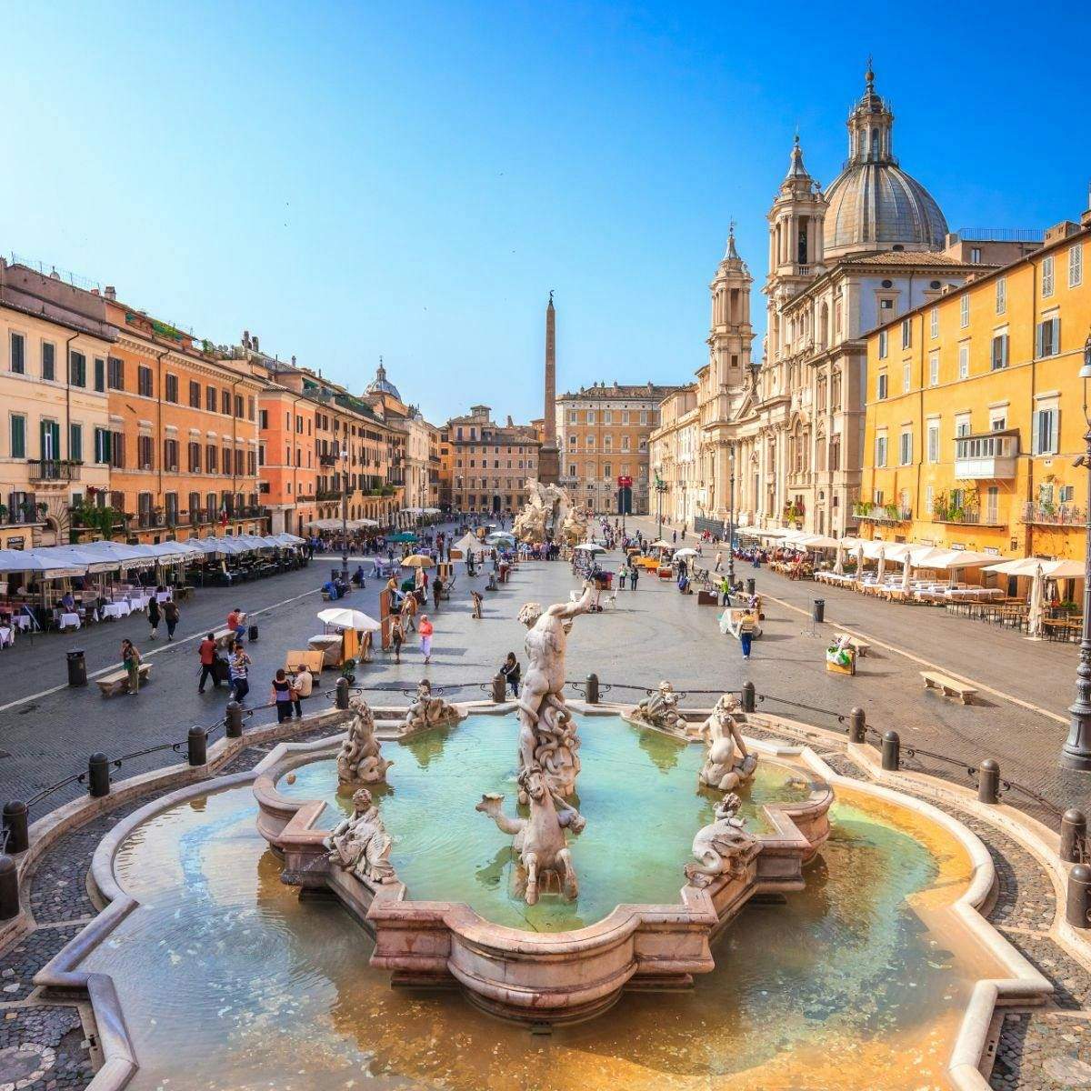 A lively square with a central fountain, surrounded by historic buildings, cafes, and people walking and sitting.