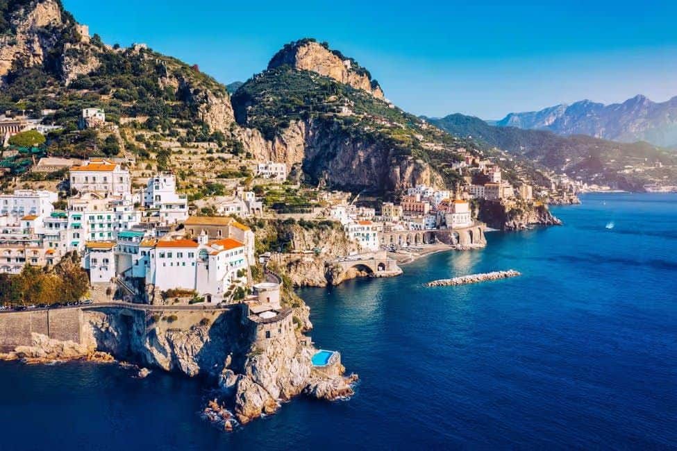 A coastal town with white buildings and red roofs on cliffs overlooking the sea, with mountains in the background.
