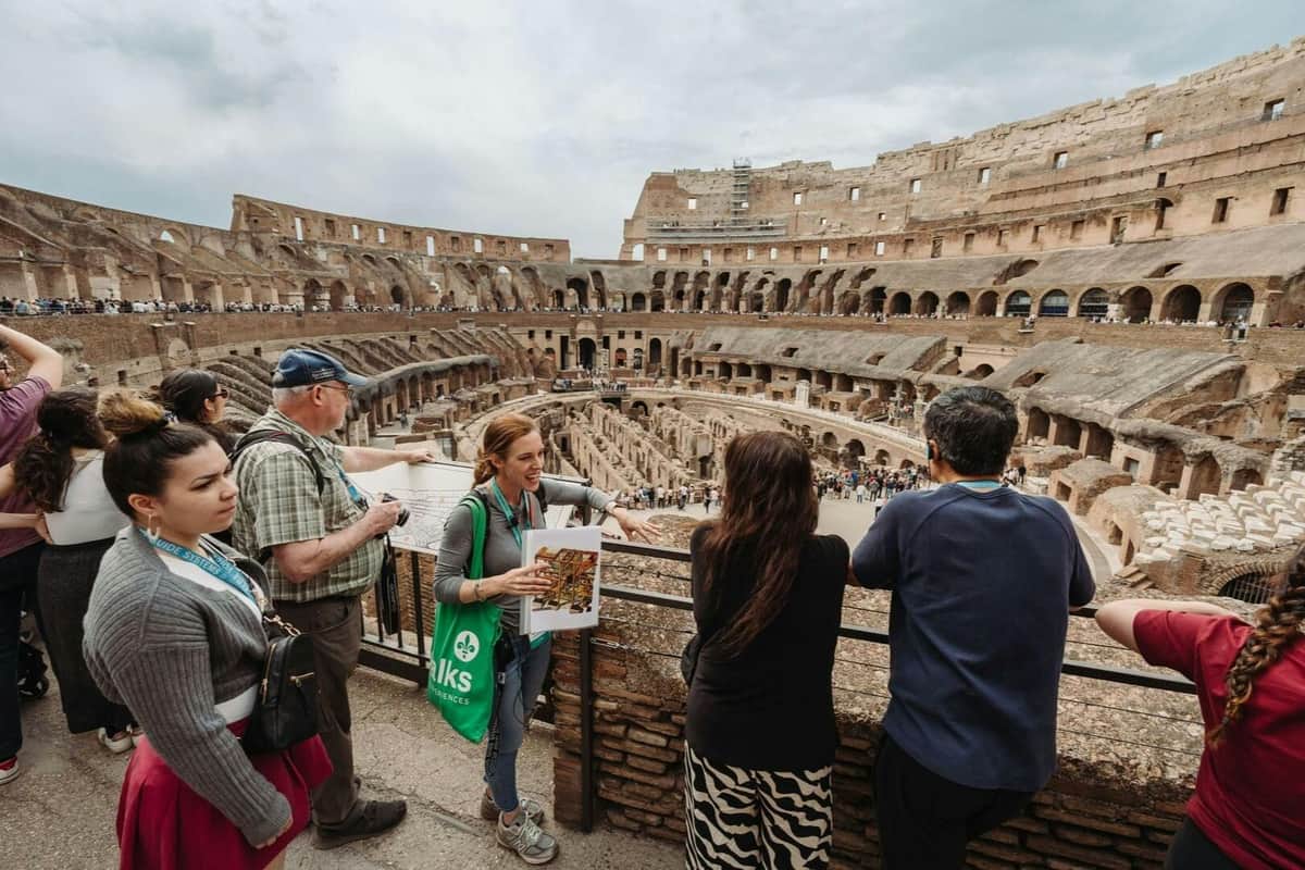 Tourists observing the interior of the Colosseum in Rome, with a tour guide explaining and holding a map.