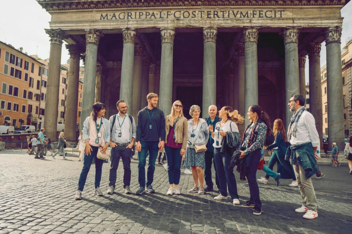 Group of tourists with audio guides standing in front of an ancient building with large columns and inscription.