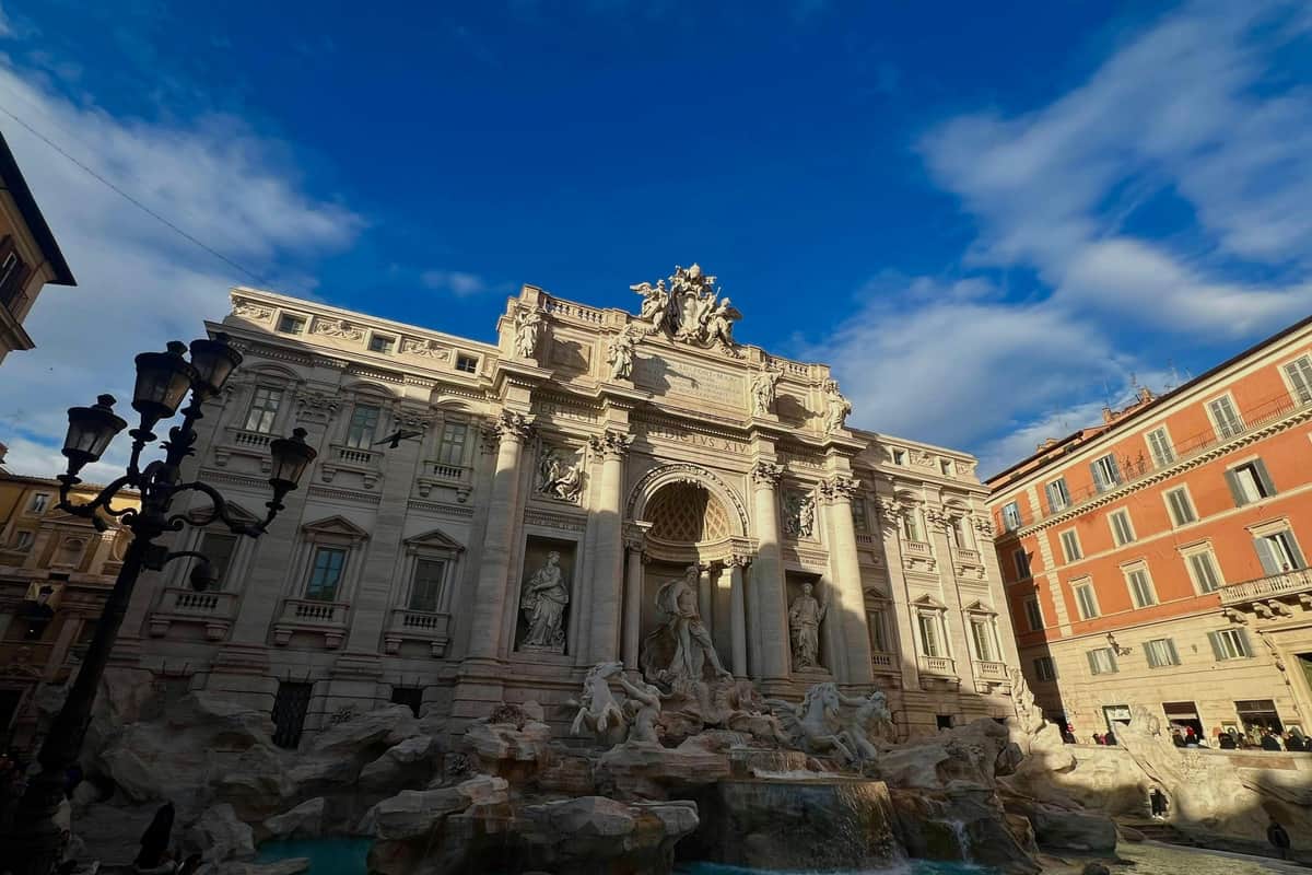 A baroque-style fountain with sculptures in a city square under a blue sky, surrounded by ornate buildings.