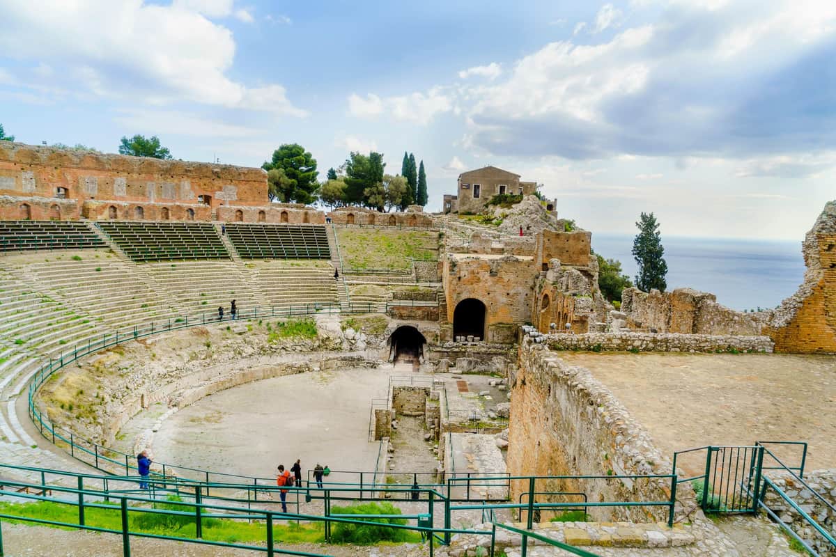 Ancient Theater of Taormina: Guided Tour Ancient amphitheater ruins with tiered seating, arched entrances, and scattered visitors, set against a backdrop of trees and cloudy sky.