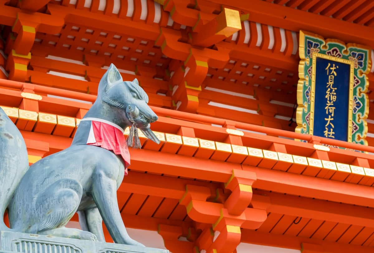 Fushimi Inari Taisha, Arashiyama, Kiyomizu-dera & Kinkaku-ji: Day Tour from Umeda with Buffet Lunch + Admission Tickets A stone fox statue with a red bib and key in its mouth, in front of an ornate wooden Japanese temple structure.
