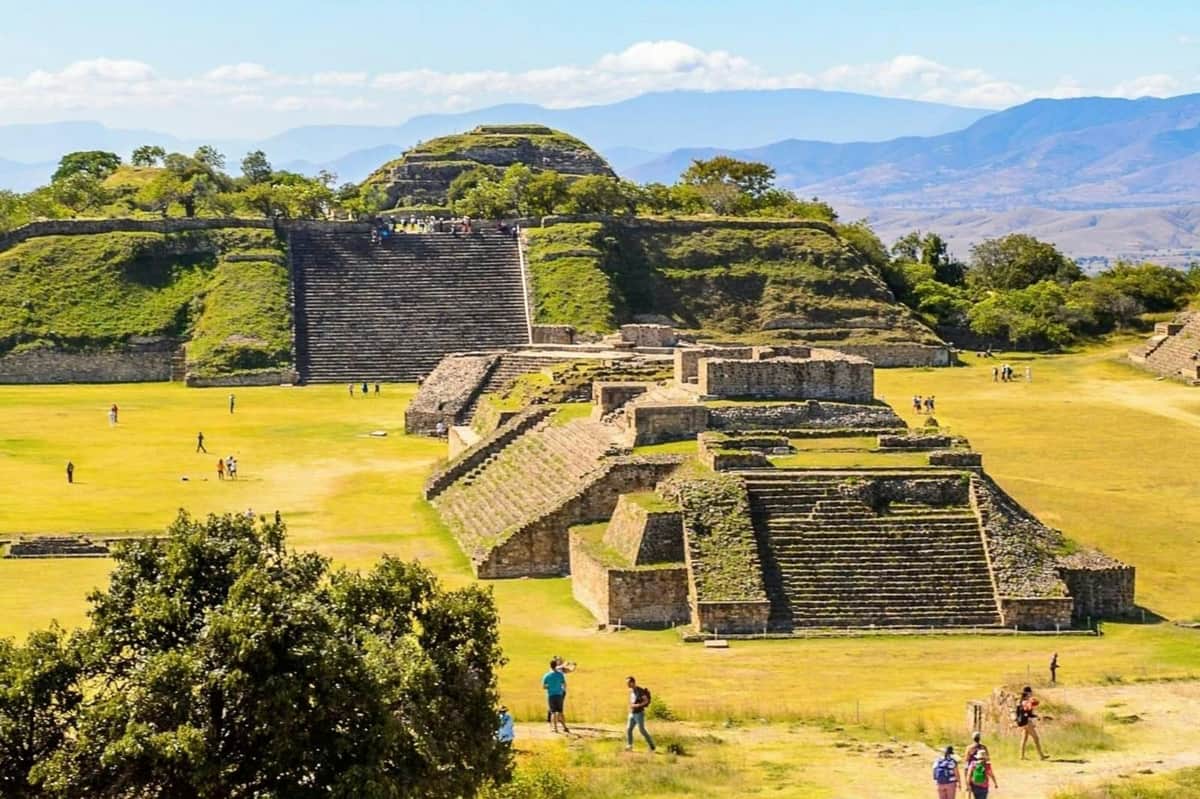 Monte Albán Archaeological Zone: Guided Tour + Transfers A scenic view of ancient pyramids and terraced structures surrounded by grassy fields and trees under a blue sky.