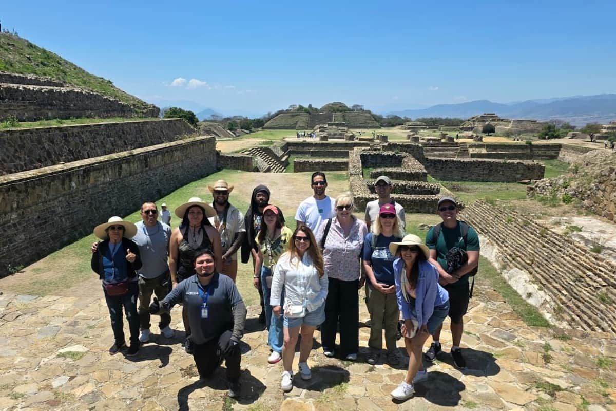 Oaxaca: Guided Tour, Transfers, Snacks + Monte Albán Entry A group of people posing together outdoors at an ancient archaeological site with stone structures and a clear blue sky.