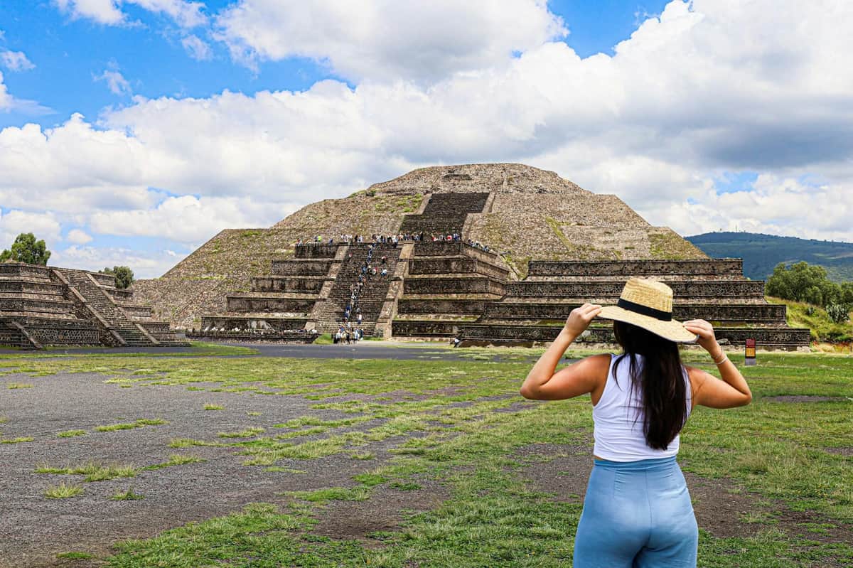 Una persona con sombrero está de pie, de cara a una gran pirámide de piedra, bajo un cielo nublado.