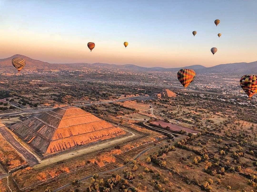 Teotihuacán Pyramids Hot Air Balloon Ride Hot air balloons float over a stepped pyramid and a sprawling landscape at sunrise or sunset, with distant mountains in the background.