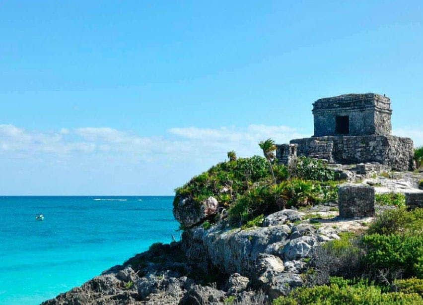 Maya Ruins of Tulum, Mariposa & Chen Ha Cenote: Guided Tour, Entry + Transport Ancient stone ruins sit atop a rocky cliff overlooking a clear blue ocean under a bright, cloud-dotted sky.