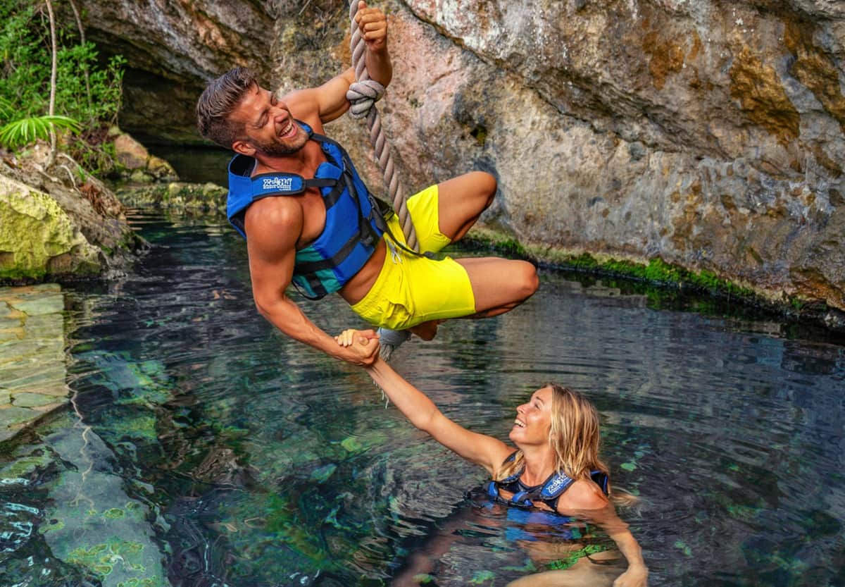 Xel-Há Park & Mayan Ruins of Tulum: Fast-Track Admission & Transport Man in yellow shorts and woman in blue swimsuit wearing life vests, smiling, and playing on a rope swing over rocky water.