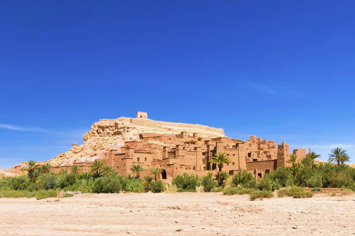 Ouarzazate: Day Tour + Aït Benhaddou Visit + Pickup from Marrakech An ancient desert fortress with mud-brick buildings and palm trees, set against a clear blue sky.