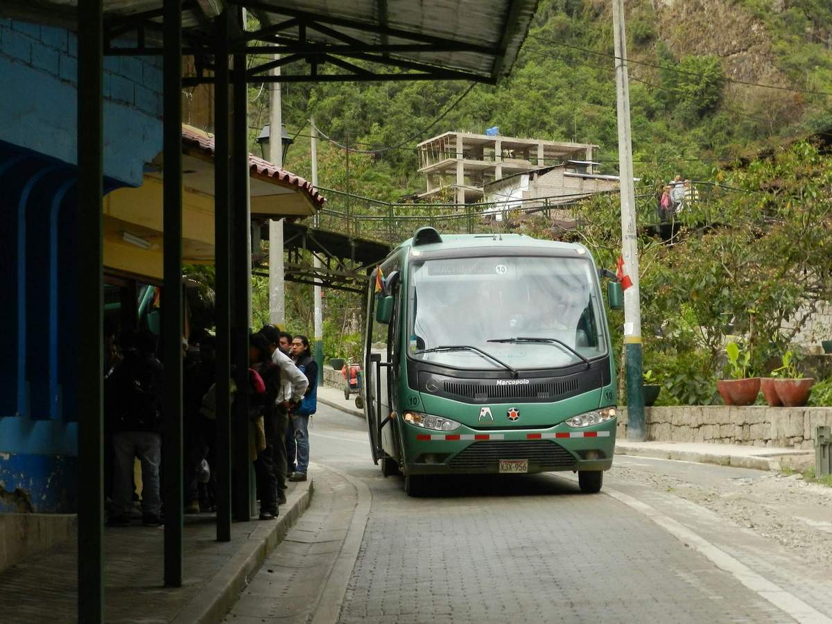 Machu Picchu: Bus from Aguas Calientes A green bus travels through a narrow, tree-lined street with people waiting at a covered stop on the left side.
