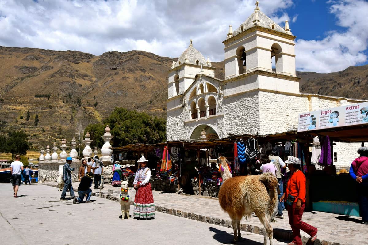Sacred Valley Day Trip from Cusco People in traditional clothing, a llama, and market stalls in front of a historic white stone church with mountains in the background.